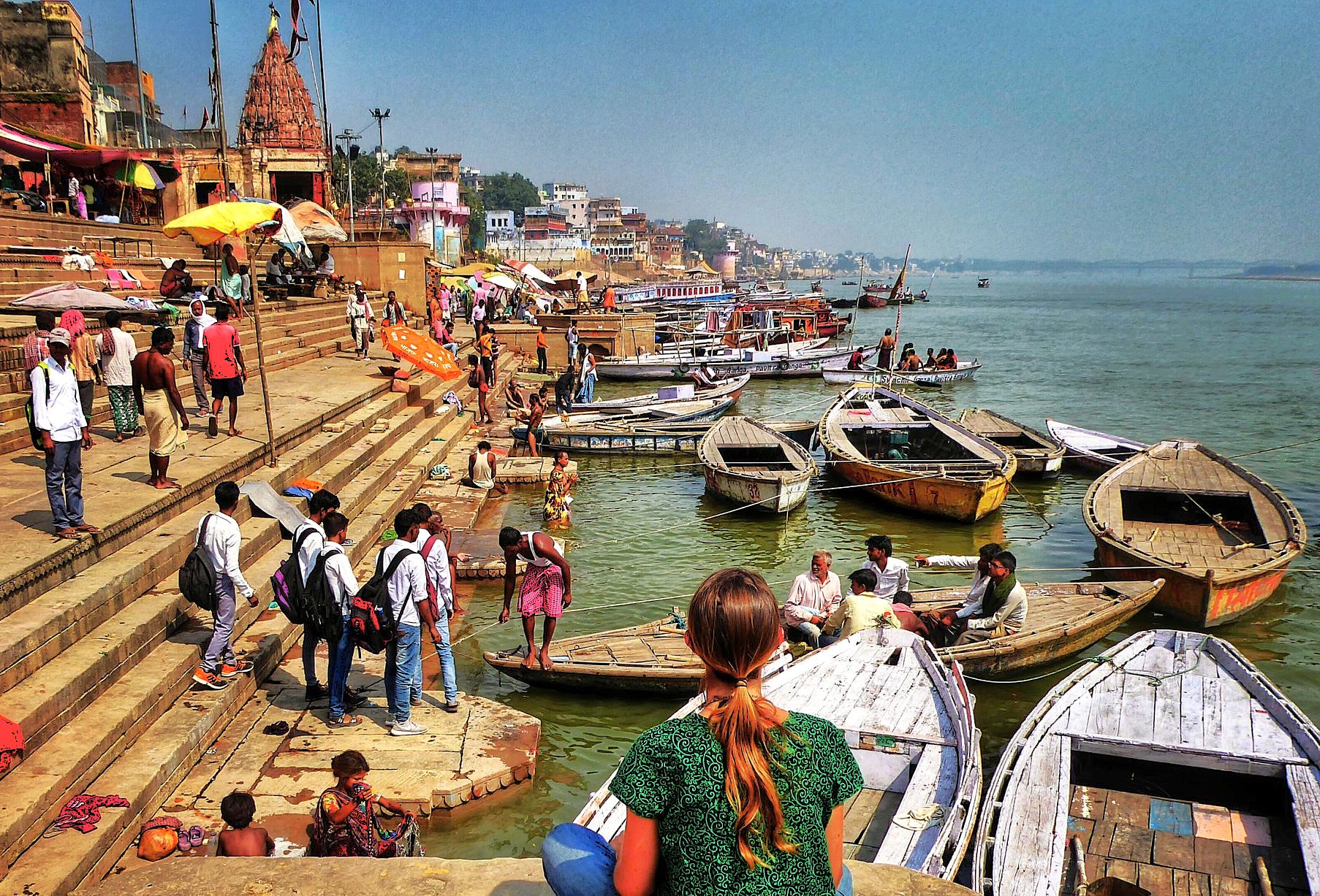 Tourists observing vibrant wooden boats moored along the Ganges ghats in Varanasi, Uttar Pradesh, India, near the revered Sanarth Temple and Buddha shrine, as locals perform ritual bathing at sunrise.