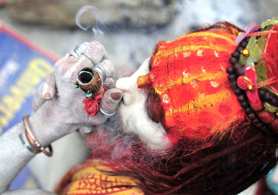 A close-up of a saffron-robed Hindu sadhu in Varanasi, Uttar Pradesh, India, near the Sanarth Temple Buddha, blowing smoke from a chillum during a morning spiritual ritual on the Ganges.