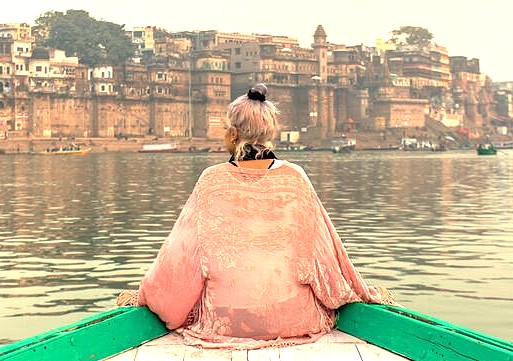 A traveler in a boat on the Ganges River in Varanasi, India, gazes at the historic ghats near Dasaswamedh Ghat, close to UNESCO World Heritage Site Sarnath, capturing spiritual serenity.