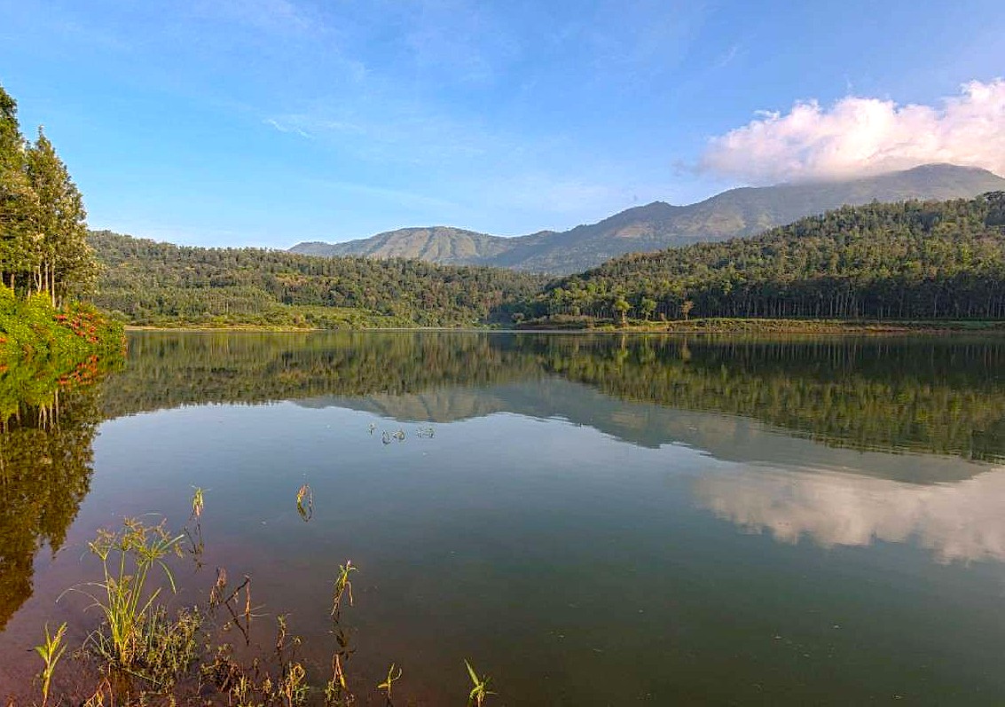 "Serene lake in the Western Ghats of southern India, calm reflective reservoir, lush forested hills, distant mountains under clear sky, eco tourism retreat, biodiversity landscape."