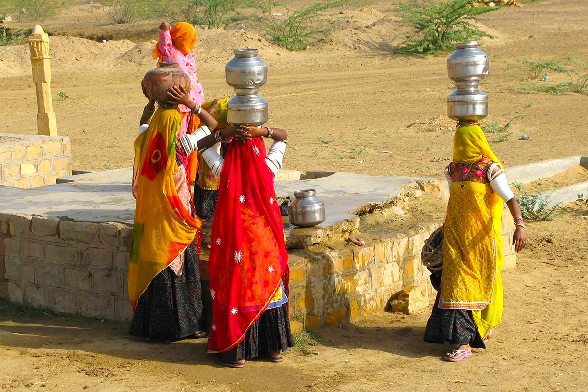 Traditional Rajasthani women in authentic vibrant attire fill water pots at a village well during the Bikaner Camel Safari near the golden sand dunes of the Thar Desert, Rajasthan, India.